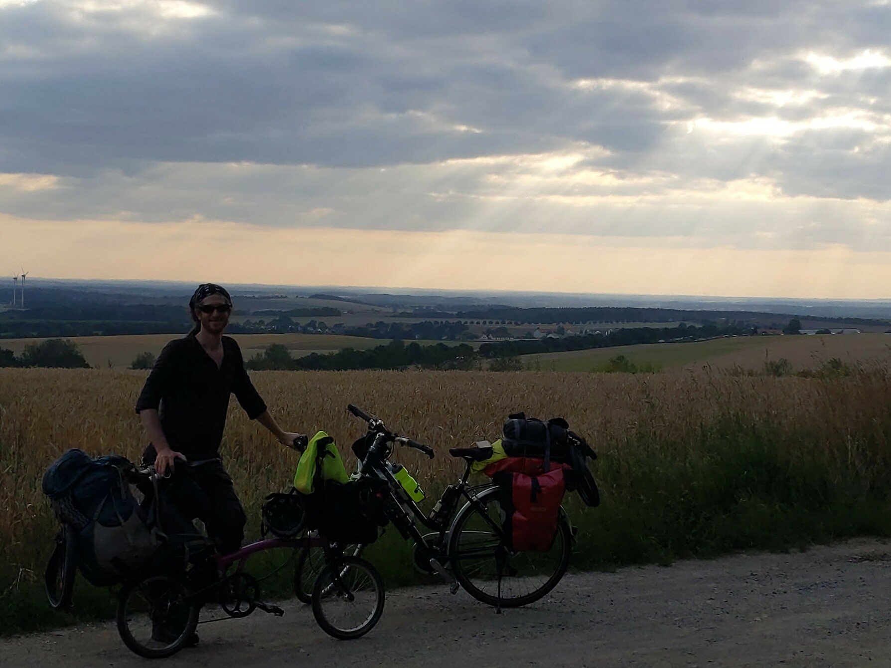 Michael Altfield standing next to a fully-loaded Brompton folding bicycle with rolling pastures of (wind) farms illuminated by crepuscular rays in the background